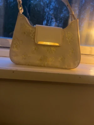 Vit axelremsväska från Anna field - Snygg vit handväska från Anna field  med axelrem och guldfärgade detaljer. Väskan har blommigt mönster i guld och en stilren rektangulär form. Perfekt accessoar för att lyfta din outfit med en trendig touch.