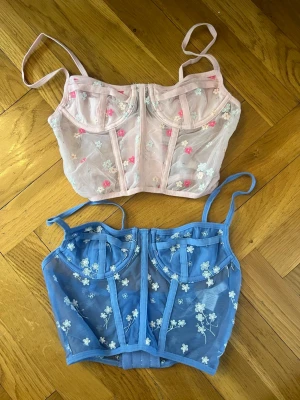 Två mesh toppar med blommor - Säljer två snygga mesh toppar med broderade blommor, en i ljusrosa och en i blått. Båda har justerbara axelband, bygel och hakknäppning bak. Perfekta för att ge outfiten en söt och trendig touch. Materialet är genomskinligt mesh med broderade detaljer. Köpta i Kalifornien i USA på Tillys. 1 för 280kr, 2 för 500kr.