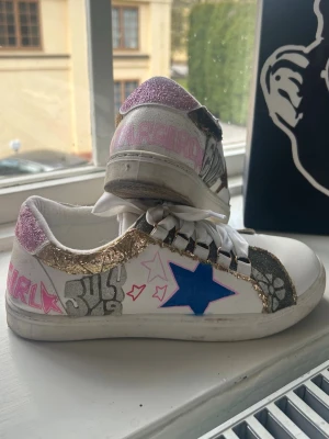 Unika egendesignade trendiga sneakers  - ❣️Köpte här på Plick av en tjej som smyckade dom själv, alltså enda paret du kommer att hitta, snyggaste jag sett så hade använt dom varje dag om de passat, därav ej använda mycket alls utan därför de ser nya ut❣️Unika vita skor med glitterdetaljer i guld och rosa, blå stjärna på sidan och handmålade motiv i silver och rosa. Skorna har vita satinsnören och coola graffiti-liknande texter och mönster. Perfekta för dig som vill sticka ut med din stil.