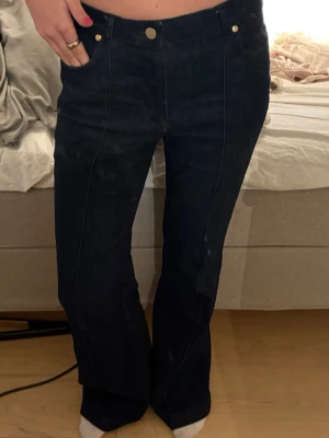 Mörkblå utsvängda jeans - Säljer ett par mörkblå jeans med utsvängda ben och markerade sömmar framtill. Klassisk femficksmodell med hög midja och silverfärgade knappar. Jeansen är i denim och har en snygg retrovibe.