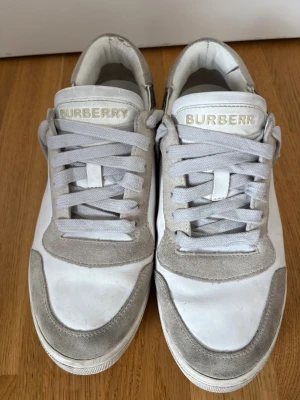 Burberry vita sneakers med rutiga detaljer - Snygga vita sneakers från Burberry med grå mockadetaljer och klassiskt rutigt mönster baktill. Skorna har vita snören och en stilren design i läder och mocka. Perfekta för dig som gillar exklusiva och trendiga sneakers med ikoniska detaljer.