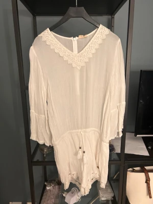 Vit strandjumpsuit med spetsdetaljer - Supersöt vit strandjumpsuit med v-ringning och spetsdetaljer runt halsen och bensluten. Lätt och luftig med 3/4-ärmar och knytband i midjan med tofsar. Perfekt att dra över bikinin på stranden. Materialet känns som tunn bomull eller viskos.