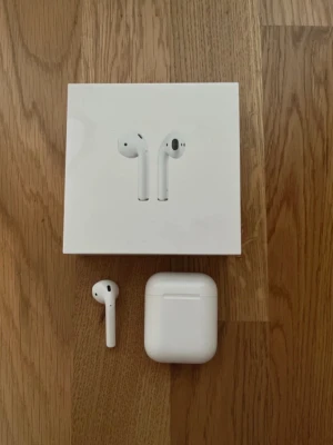 Apple AirPods med laddningsetui - Trådlösa Apple AirPods i klassisk vit färg med laddningsetui. Snygg och stilren design i plast, perfekt för dig som vill ha smidiga hörlurar till mobilen. Kommer med originalförpackning. Laddare ingår ej. Har tappat bor ena men funkar bra ändå