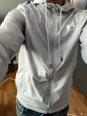 Ljusgrå vindjacka från Under Armour - Snygg ljusgrå vindjacka från Under Armour med dragkedja framtill och justerbar huva med snören. Jackan har en sportig look och är tillverkad i ett lätt material som passar perfekt för blåsiga dagar. Logotypen syns tydligt på bröstet. Dock har den ett märke som inte försvinner 