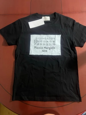 Maison Margiela svart t-shirt - Svart t-shirt från Maison Margiela med klassisk vit patch framtill med siffror och logotyp. T-shirten har rund halsringning och korta ärmar. Materialet är mjuk bomull som känns skön mot huden. Perfekt för dig som gillar stilrena och trendiga plagg. OBS! Tröjan är lite tightare runt bröstet och kan därmed även passa som S
