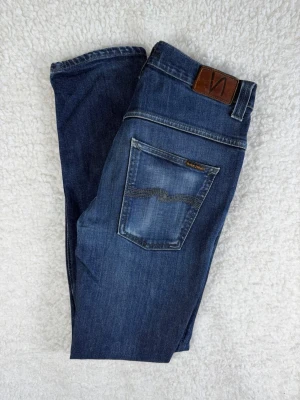Nudie jeans Thinn Finn (Dry Dark Grey) W33 L34 - Snygga mörkblå jeans från Nudie Jeans med klassisk femficksdesign och bruna detaljer på bakfickan. Jeansen har en smal passform och är tillverkade i bomull med lätt stretch för extra komfort. Perfekta för en avslappnad och trendig look.