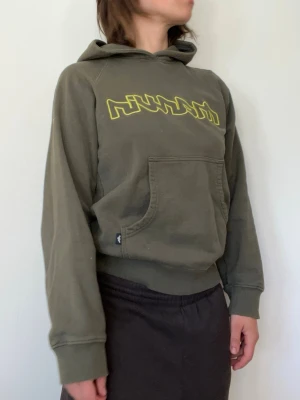 Olivgrön hoodie från Marvin  - Snygg olivgrön hoodie från Marvin med gul grafisk text på bröstet. Hoodien har en klassisk känguruficka framtill och ribbade muddar vid ärmslut och nederkant. 