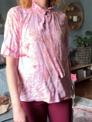 Rosa mönstrad blus med knytband - Säljer en luftig rosa blus med korta ärmar och ett unikt mönster i olika rosa nyanser. Blusen har ett dekorativt knytband vid halsen och en avslappnad passform. Perfekt för dig som gillar en färgglad och bekväm stil. 100% viskos supermjuk