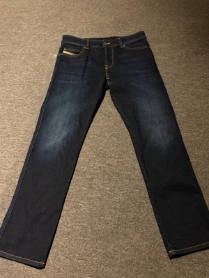 Mörkblå raka jeans från Diesel - Snygga mörkblå jeans från Diesel med raka ben och klassiska kontrastsömmar. Jeansen har fem fickor och en liten logga på myntfickan. Perfekta för en stilren och avslappnad look. Storlek 29,30