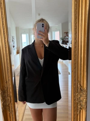 Svart oversized kavaj - Stilren svart oversized kavaj med klassisk krage och enkelknäppning. Kavajen har raka axlar, två fickor framtill och är tillverkad i ett slätt tyg som ger en snygg och modern look. Perfekt att slänga över en topp eller klänning för en trendig outfit.