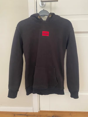 Svart hoodie från Hugo Boss - Svart hoodie från Hugo Boss med röd logotyp på bröstet. Klassisk modell med huva, känguruficka och ribbade muddar vid ärmslut och nederkant. Perfekt för en avslappnad och stilren look.