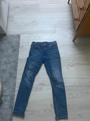 Blå jeans från Tommy Hilfiger - Jeans från Tommy Hilfiger. Jag säljer ett par blåa Tommy Hilfiger jeans då de blivit för små. Passformen är ganska tight och lite avsmalnande nertill. Några stygn till läder patchen i bak har gått upp.