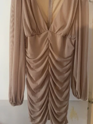 Beige omlottklänning PrettyLittleThing - Säljer en beige kortklänning från PrettyLittleThing med omlottdesign, djup v-ringning och långa puffärmar. Klänningen har snygg rynkning framtill som ger en cool och trendig look. Perfekt för dig som vill sticka ut lite extra!