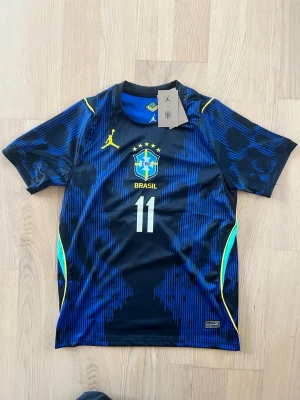 Blå Brasilien Raphinia fotbollströja Jordan VM 2026 - Snygg blå och svart Brasilien fotbollströja från Jordan med nummer 11 på bröstet. Tröjan har gula detaljer, CBF-logga och korta ärmar. Perfekt för fotbollsträning eller att bära som supporter. Materialet är lätt och andas.