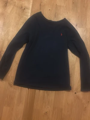 Mörkblå långärmad tröja Polo Ralph Lauren - Mörkblå långärmad tröja från Polo Ralph Lauren med klassisk röd logga broderad på bröstet. Enkel och stilren design med rund halsringning. Perfekt för en avslappnad och snygg look.