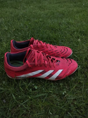 Adidas Predator röda fotbollsskor - Säljer ett par röda Adidas Predator fotbollsskor med vita detaljer och klassiska tre ränder på sidan. Skorna har snörning, låg profil och mönstrad ovandel för bättre bollkontroll. Perfekta för dig som vill sticka ut på planen.
