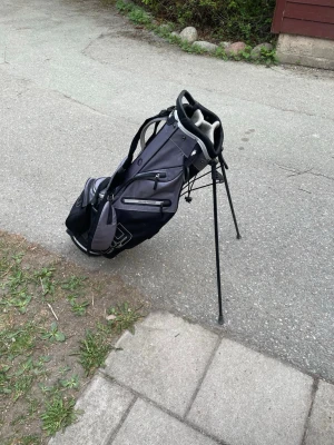 Dry golf bag  - Golfbag i svart och grått med praktiskt benstativ för enkel uppställning på banan. Flera fack för klubbor och tillbehör samt bärrem för smidig transport. Tillverkad i slitstarkt tygmaterial och har förstärkt handtag upptill.