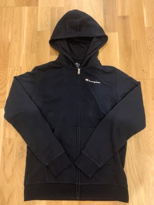 Svart zip-hoodie från Champion - Svart hoodie med dragkedja från Champion. Klassisk design med huva och fickor framtill. Champion-loggan är broderad på bröstet i vitt och rött. Mjuk bomullskänsla och ribbade muddar vid ärmslut och nederkant. Perfekt för en avslappnad streetstyle. Passar bäst för dig runt 154 ish. Kan bara mötas, förlåt.