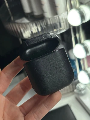 Svart Disney AirPods-fodral - Snyggt svart AirPods-fodral i läderimitation med diskret Mickey Mouse-mönster. Fodralet har en stilren look och är perfekt för dig som vill skydda dina hörlurar med lite extra Disney-vibe. Passar AirPods och har en praktisk ögla på baksidan.