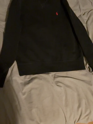 Svart sweatshirt från Ralph Lauren - En svart sweatshirt från Ralph Lauren med klassisk rund hals och lång ärm. Tröjan har en liten röd broderad logga på bröstet och breda muddar vid ärmslut och nederkant. Perfekt för en avslappnad och stilren look.
