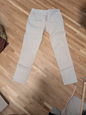 Beiga chinos med rak passform - Snygga beiga chinos med rak passform och elastisk midja. Byxorna har klassisk design utan synliga detaljer och är tillverkade i ett mjukt, lätt tyg som känns bekvämt hela dagen. Perfekta för en stilren och avslappnad look.