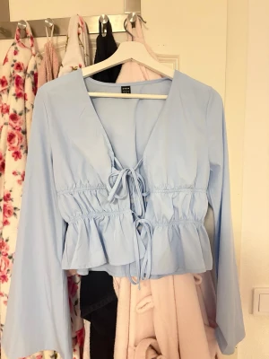 Ljusblå blus med knytning fram - Trendig ljusblå blus från NA-KD med v-ringning och två dekorativa knytband framtill. Blusen har volang nedtill och långa ärmar. Tillverkad i ett lätt, vävt material som ger en luftig känsla.