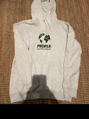Grå hoodie People LoveYourHood - Snygg grå hoodie från People LoveYourHood med svart tryck av en världskarta och texten 'PEOPLE LoveYourHood' på bröstet. Klassisk modell med huva och dragsko samt ribbade muddar vid ärmar och nederkant. Perfekt för en avslappnad streetwear-look.
