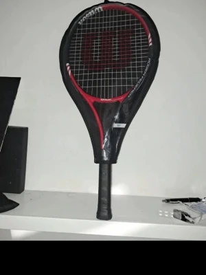 Wilson tennisracket svart/röd - Tennisracket från Wilson i svart och rött med tydlig logga på strängarna. Handtaget är svart och har Wilson-tryck. Racketen har en klassisk form och är tillverkad i slitstarkt material, perfekt för dig som gillar att spela tennis.