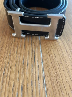 Svart skärp med stor H-spänne - Snyggt svart bälte med en stor, silvrig H-formad metallspänne och vita kontrastsömmar. Perfekt accessoar för att lyfta vilken outfit som helst. Klassisk och stilren design.