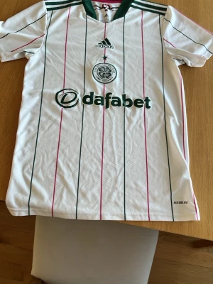 Vit Celtic FC matchtröja Adidas - Snygg vit matchtröja från Celtic FC med gröna och rosa ränder, Adidas-logga och klubbmärke på bröstet. Sponsorloggor från Dafabet och Magners. Tillverkad i lätt och ventilerande AEROREADY-material, perfekt för träning eller match.
