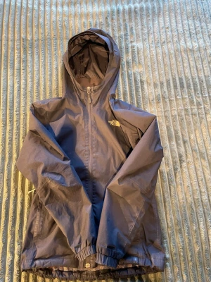 Svart regnjacka från The North Face - Snygg svart regnjacka från The North Face med huva och dragkedja framtill. Jackan har elastiska muddar vid ärmslut och nederkant samt två sidofickor. Perfekt för regniga dagar och enkel att packa ner i väskan. Tillverkad i vattenavvisande material.