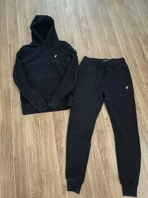 Svart loungewear-set från Lyle & Scott - Mysigt svart loungewear-set från Lyle & Scott med matchande hoodie och byxor. Setet har gul broderad logga på både tröja och byxa. Hoodien har känguruficka och dragsko i huvan, byxorna har resår i midjan och muddar vid benslut. Perfekt för chill dagar hemma. Storlek XS! Skriv till mig vid intresse eller frågor!