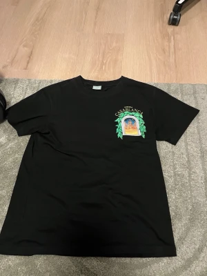 Svart Casablanca t-shirt med tryck - Snygg svart t-shirt från Casablanca med färgglatt tryck av ett fönster och gröna blad på bröstet och ryggen. Texten 'Avenida Casablanca' syns tydligt. Klassisk passform och rund hals. Perfekt för dig som vill sticka ut med en unik design.