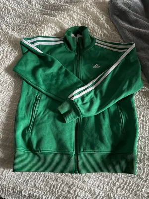Grön Adidas ziptröja med vita ränder - Säljer en grön ziptröja från Adidas med klassiska vita ränder längs ärmarna och Adidas-logga på bröstet. Tröjan har hög krage, dragkedja framtill och två fickor med dragkedja. Ribbstickade muddar vid ärmslut och nederkant. Perfekt för en sportig look.