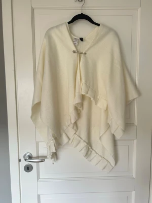 Krämvit ponchotröja från H&M - Krämvit ponchotröja från H&M med volangkant och dekorativ säkerhetsnål framtill. Tröjan har en lös och flowy passform och är gjord i ett mjukt tyg som känns skönt mot huden. Perfekt att slänga över en outfit för extra stil.