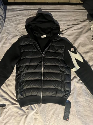 Svart pufferjacka med huva från Moncler - Snygg svart cardigan från Moncler med huva och dragkedja framtill. Jackan har stickade ärmar, två fickor med dragkedja och en vit M-logga på ena ärmen. Klassisk Moncler-logga på ärmen och quiltad front i blankt material. Byte är också intressant 