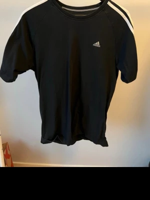 Svart Adidas t-shirt med vita ränder - Snygg svart t-shirt från Adidas med klassiska vita ränder på axlarna och liten Adidas-logga på bröstet. T-shirten har rund hals och korta ärmar, perfekt för en sportig och avslappnad stil. Tillverkad i mjuk bomull för skön känsla.