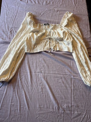 Vit croppad blus med snörning - Trendig ljusbeige croppad blus med långa puffärmar och snygg snörning framtill. Blusen har en fyrkantig ringning och är tillverkad i ett lätt, krinklat bomullstyg som ger en avslappnad men chic vibe.