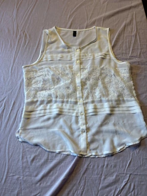 Vit ärmlös blus med spetsdetaljer - Säljer en vit ärmlös blus med knappar framtill och transparenta spetsdetaljer över bröstet. Blusen har en lätt och luftig känsla, perfekt för varma dagar. Snygga horisontella sömmar ger extra stil. Passar till både jeans och kjol.