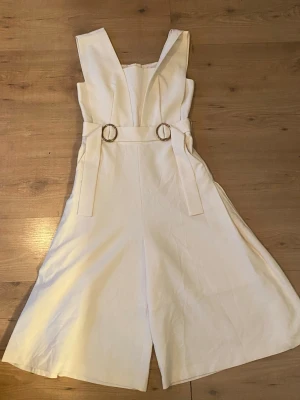 Vit ärmlös jumpsuit med ringdetaljer - Säljer en snygg vit jumpsuit med breda axelband och vida ben. Jumpsuiten har två dekorativa ringdetaljer i midjan och är minimalistiskt. Materialet känns mjukt och har en lätt struktur. Helt oanvänd 🤗
