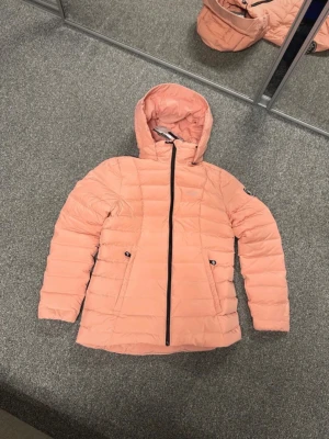 Ljusrosa dunjacka från Pelle P - Snygg ljusrosa dunjacka från Pelle P med huva och svarta dragkedjor. Jackan har två fickor framtill och quiltad design som ger en modern look. Perfekt för kalla vinterdagar och riktigt skön att bära. Märkeslogga på bröstet.