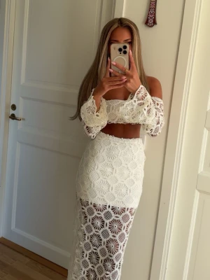 Vit virkad tvådelad set med lång kjol - Säljer ett vitt tvådelat set med virkad design. Toppen har långa ärmar och off-shoulder-modell, medan kjolen är lång och genombruten med blommigt mönster. Setet är gjort i ett mjukt, virkat material som ger en bohemisk känsla. Perfekt för dig som gillar unika och trendiga plagg.