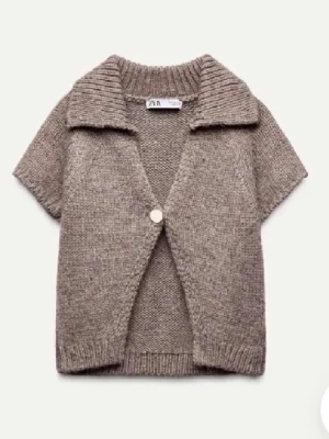 Beige stickad kofta från Zara - Kortärmad stickad kofta i beige från Zara med bred krage och en knapp framtill. Koftan har en avslappnad passform och ribbade detaljer vid ärmslut, nederkant och krage. Perfekt att slänga över en t-shirt för en chill look.