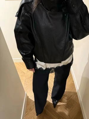 Svart oversized skinnjacka - Cool svart skinnjacka med oversized passform och höga axlar. Jackan har en unik asymmetrisk knäppning och högt krage med knappar. Perfekt för dig som vill ha en edgy och trendig look. Väldigt populär just nu och denna är helt oanvänd med lappen kvar. 