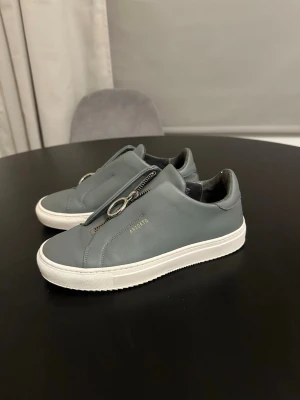 Gråa sneakers från Axel Arigato - Snygga grå sneakers från Axel Arigato i läder med vita sulor och coola dragkedjor med ringdetaljer istället för klassiska snören. Minimalistisk design med diskret logga i guld på sidan. Perfekta för en clean och modern stil.