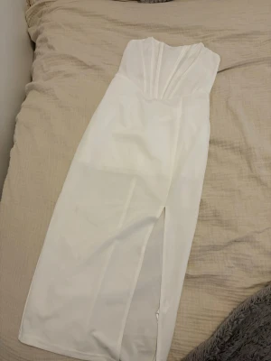 Vit axelbandslös långklänning - Snygg vit axelbandslös långklänning med draperad överdel och slits framtill. Perfekt för dig som vill ha en elegant och stilren look. Klänningen har en figurnära passform och är tillverkad i ett mjukt material. Endast använd en gång och inga fläckar på klänningen. Kom med egna bud och hör av er vid intresse 