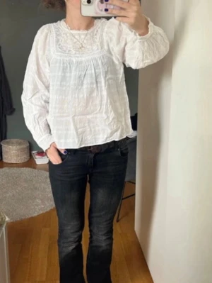 Vit boho-blus med spetsdetaljer - Söt vit blus i boho-stil med broderade spetsdetaljer upptill och rynkade långärmar. Tillverkad i ett lätt, luftigt tyg som ger en avslappnad och trendig look. Perfekt att matcha med jeans eller kjol för en chill vibe.