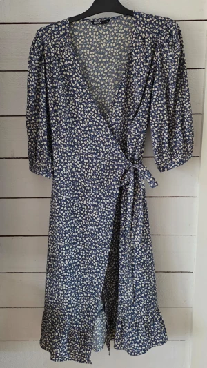 Blommig omlottklänning från ONLY - Supersöt blå omlottklänning från ONLY med vitt blommönster, knyt i midjan och volangkant nertill. Klänningen har v-ringning och halvlånga puffärmar. Perfekt för sommarens alla tillfällen!