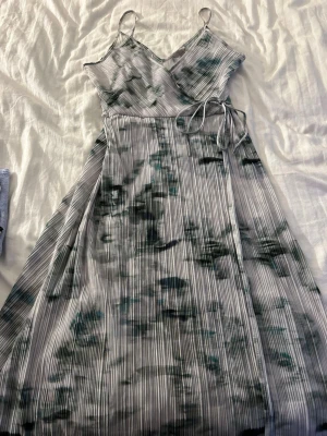 Lång omlottklänning med knytband - Säljer en lång omlottklänning med smala axelband och knyt i midjan. Klänningen har ett randigt mönster i vitt och ljusgrönt med inslag av mörkgrönt. Materialet känns lätt och flowy, perfekt för varma dagar. Snygg v-ringning och avslappnad passform.