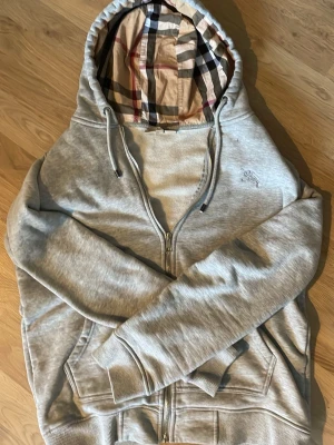 Grå zip hoodie från Burberry - Säljer en grå zip hoodie från Burberry med klassiskt rutigt foder i huvan och dragsko. Hoodien har fickor framtill, ribbade muddar och en liten broderad logga på bröstet. Perfekt för en avslappnad och stilren look. Hoodien är i perfekt form med absolut inga slitage eller märken. Lägger ut för ett bra pris ocks👍